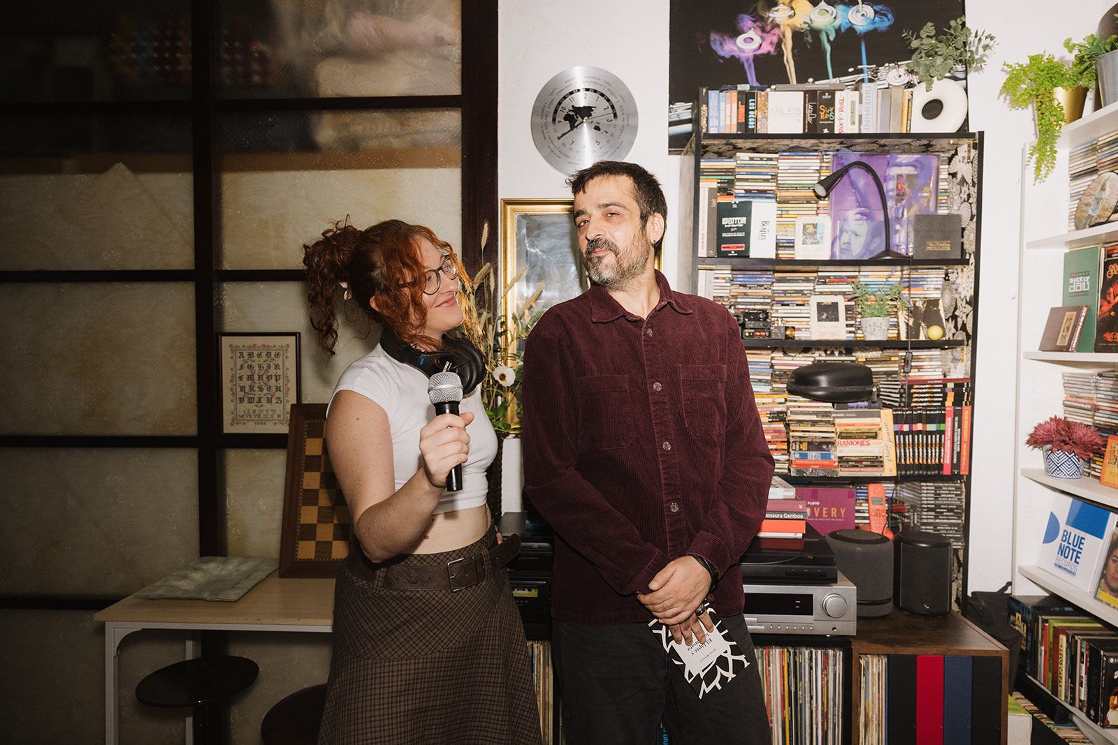 Núria con un micrófono y Toni a su lado, sonriendo, en una sala llena de discos y libros